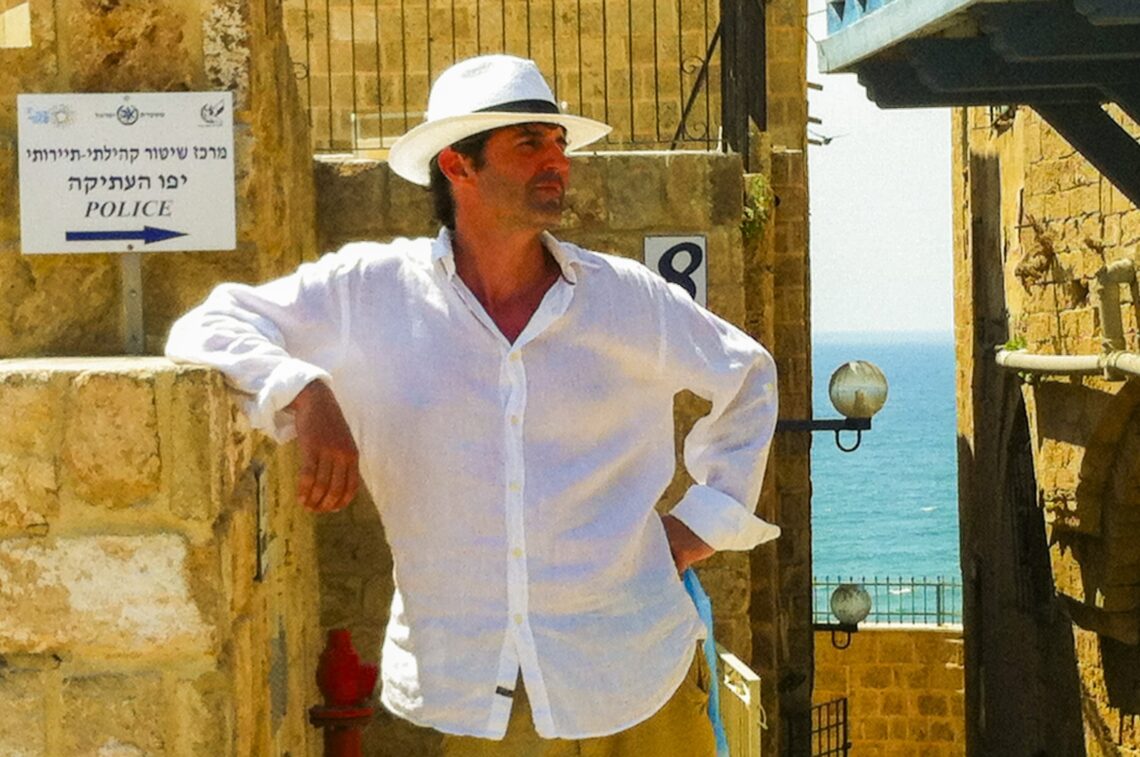 Roger Stein im weißen Hemd und Panamahut lehnt in einer Gasse der Altstadt von Jaffa, im Hintergrund das Meer