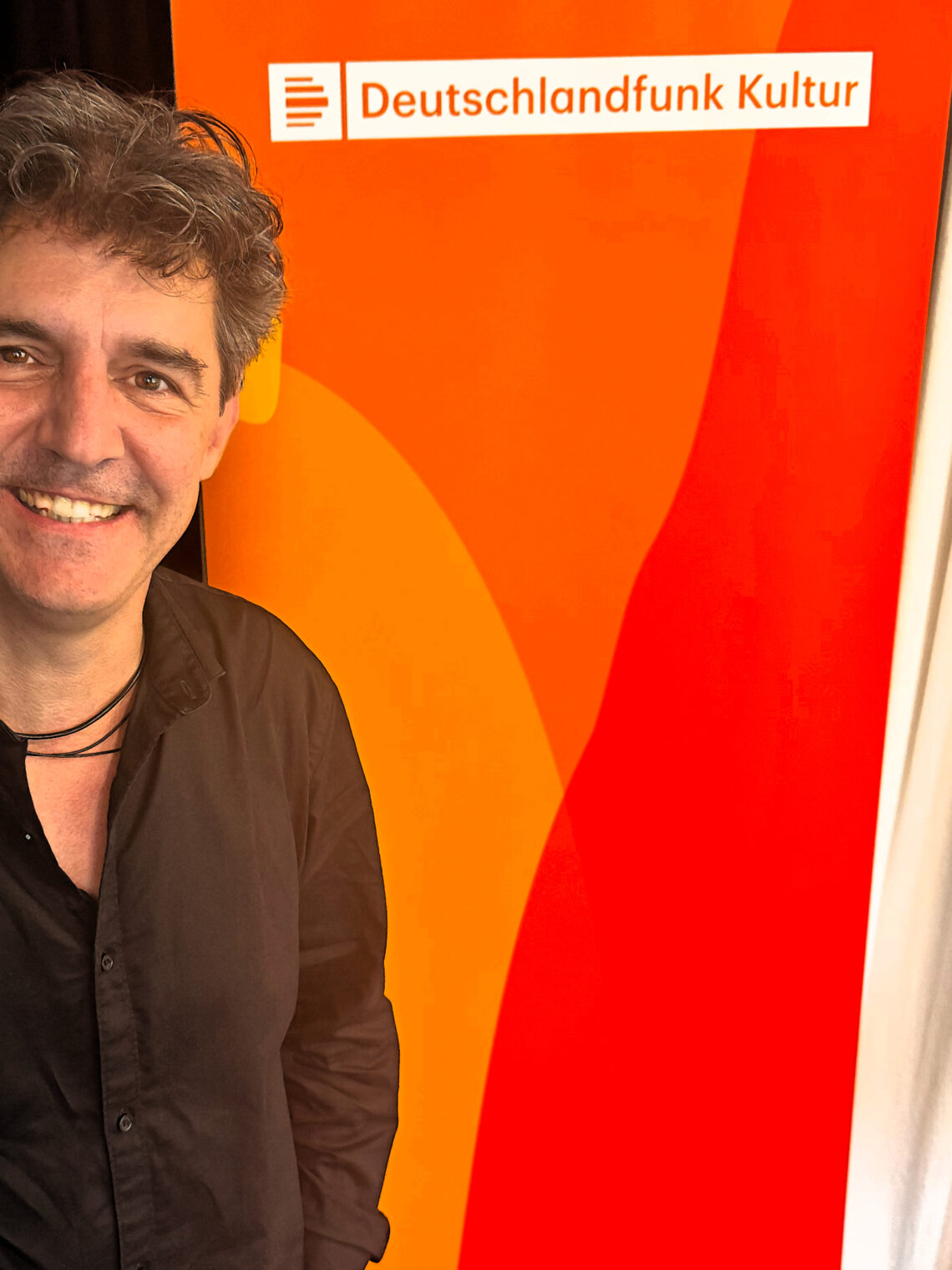 Roger Stein lächelnd vor dem Banner von Deutschlandfunk Kultur im Funkhaus Berlin