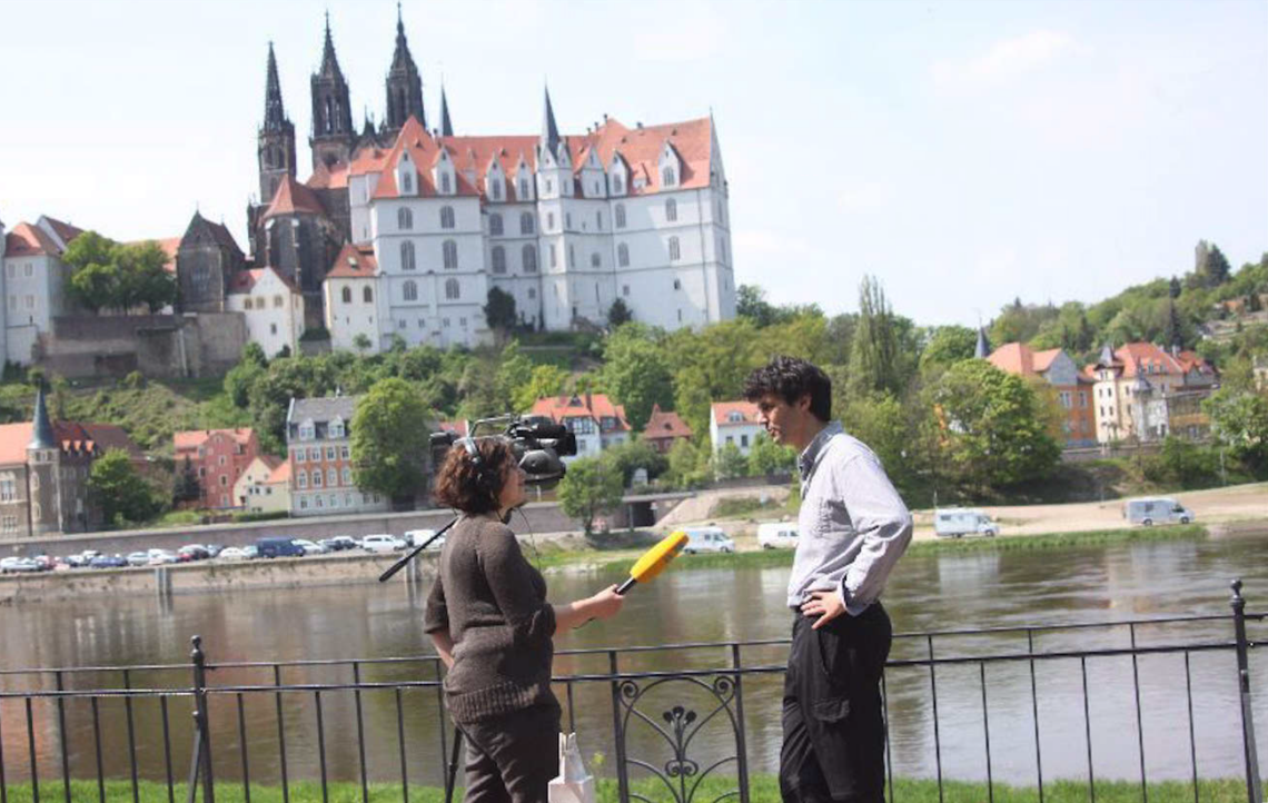 Roger Stein im Gespräch mit einer Reporterin an der Elbe, im Hintergrund die Albrechtsburg Meißen