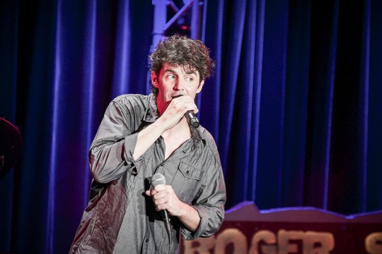 Roger Stein im grauen Hemd singt mit zwei Handmikrofonen vor blauem Bühnenvorhang
