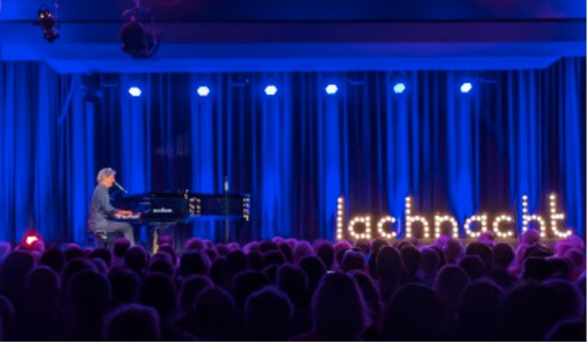 Roger Stein am Flügel auf der blau beleuchteten Bühne der Stadthalle Montabaur, daneben der Leuchtschriftzug „lachnacht“, im Vordergrund das Publikum