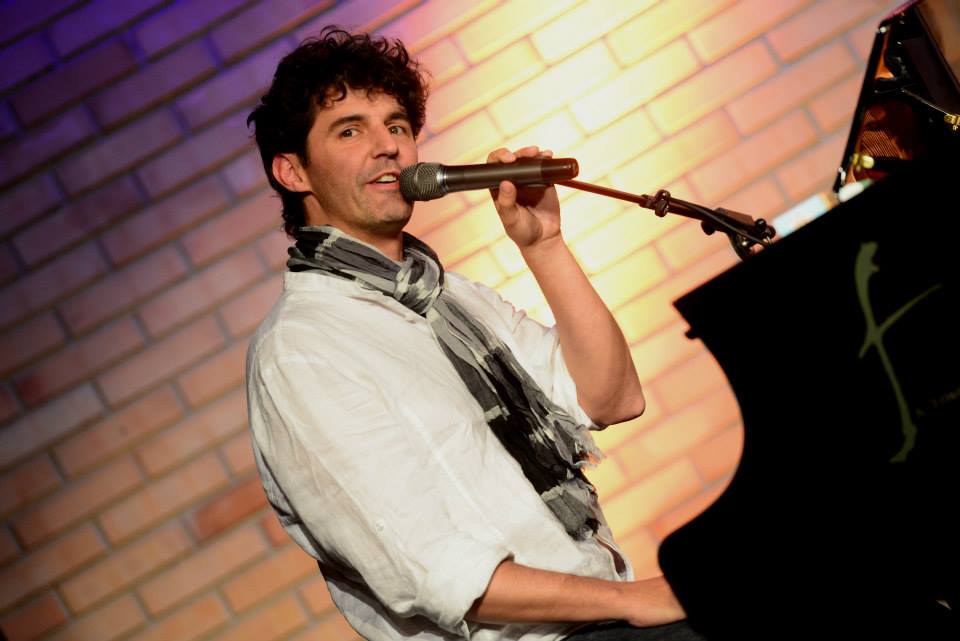 Roger Stein im weißen Hemd mit Schal singt am Flügel vor einer beleuchteten Backsteinwand