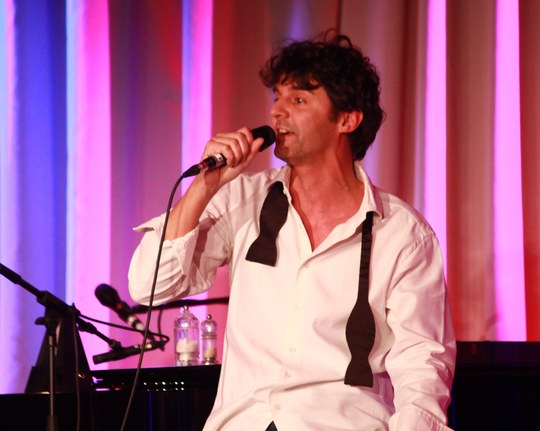 Roger Stein im weißen Hemd mit offener Fliege singt ins Mikrofon vor bunt beleuchtetem Vorhang