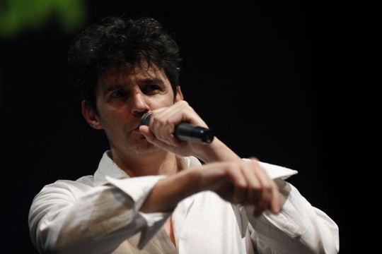Roger Stein im weißen Hemd singt mit Handmikrofon vor dunklem Hintergrund