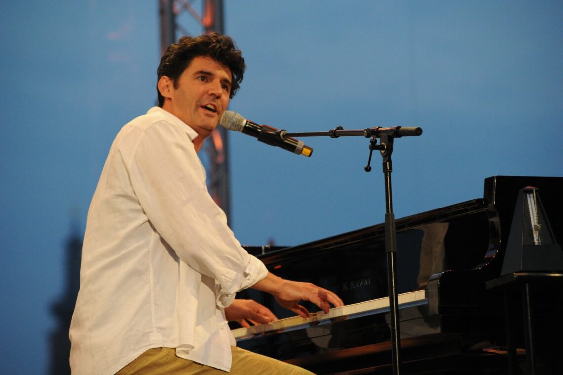 Roger Stein im weissen Hemd singt am Flügel ins Mikrofon, Open-Air-Bühne vor blauem Himmel
