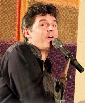 Roger Stein singt am Mikrofon vor einem bunten Wandteppich