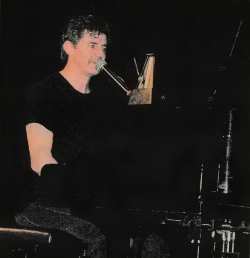 Roger Stein im schwarzen T-Shirt am Flügel mit Mikrofon und Metronom, dunkler Bühnenhintergrund