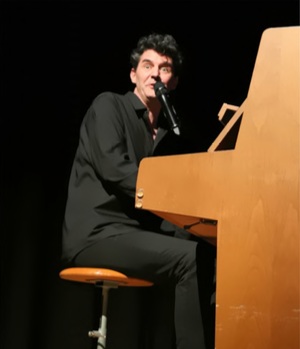 Roger Stein im schwarzen Hemd am hellen Holzklavier mit Mikrofon, vor schwarzem Hintergrund