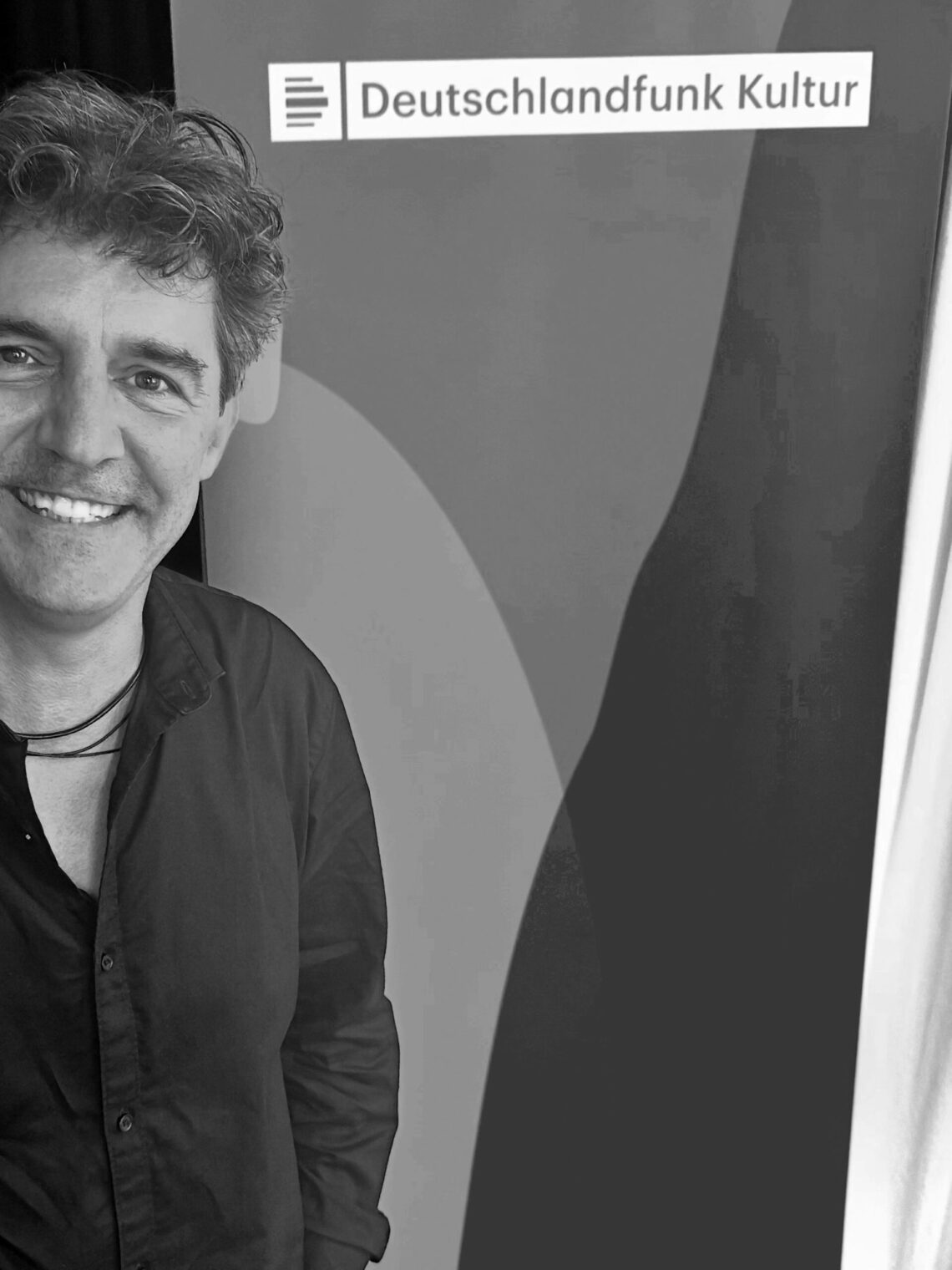Roger Stein lächelnd vor dem Banner von Deutschlandfunk Kultur im Funkhaus Berlin, Schwarzweissfoto